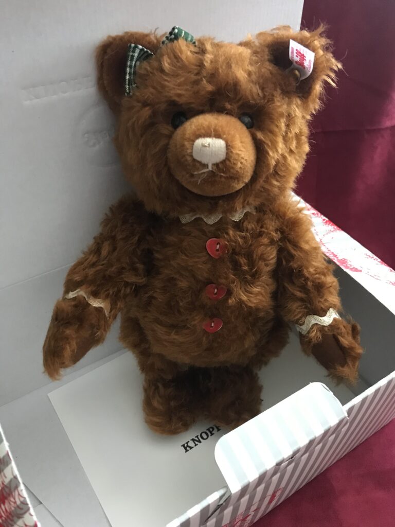 Rotbrauner Steiffteddy Gingerbread mit karierter Schleife im rechten und Ohrfahne im linekn Ohr und roten Herzknöpfen im Karton stehend