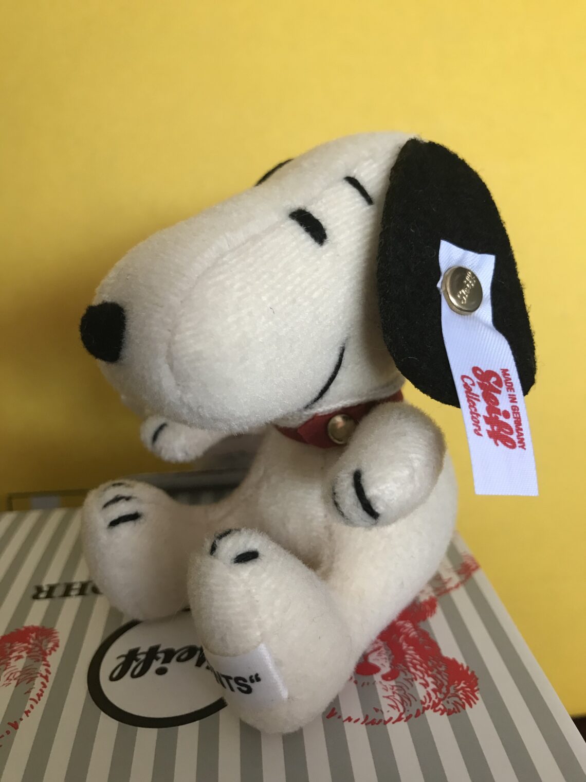 Luxusplüsch.Steiff Beagle Snoopy in weiß mit schwarzen Ohren und schwarz bestickten Details sowie weißer Ohrfahne mit rotem Steiff Logo-Schriftzug sitzend auf Steiff Karton vor gelbem Hintergrund