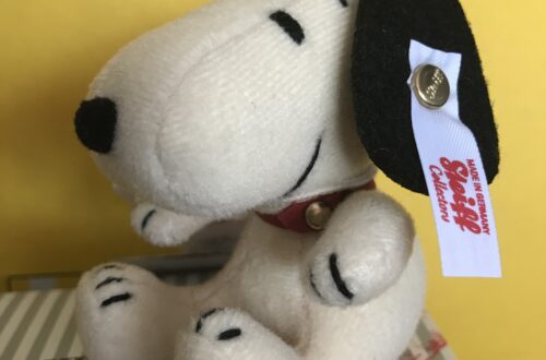 Luxusplüsch.Steiff Beagle Snoopy in weiß mit schwarzen Ohren und schwarz bestickten Details sowie weißer Ohrfahne mit rotem Steiff Logo-Schriftzug sitzend auf Steiff Karton vor gelbem Hintergrund