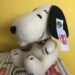 Luxusplüsch.Steiff Beagle Snoopy in weiß mit schwarzen Ohren und schwarz bestickten Details sowie weißer Ohrfahne mit rotem Steiff Logo-Schriftzug sitzend auf Steiff Karton vor gelbem Hintergrund
