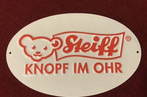 Weißes Blechschild mit hellrotem Steiff Logo mit Teddy und Textfahne Steiff und Text darunter "Knopf im Ohr"