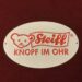 Weißes Blechschild mit hellrotem Steiff Logo mit Teddy und Textfahne Steiff und Text darunter "Knopf im Ohr"