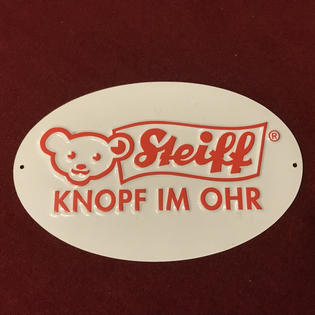 Weißes Blechschild mit hellrotem Steiff Logo mit Teddy und Textfahne Steiff und Text darunter "Knopf im Ohr"